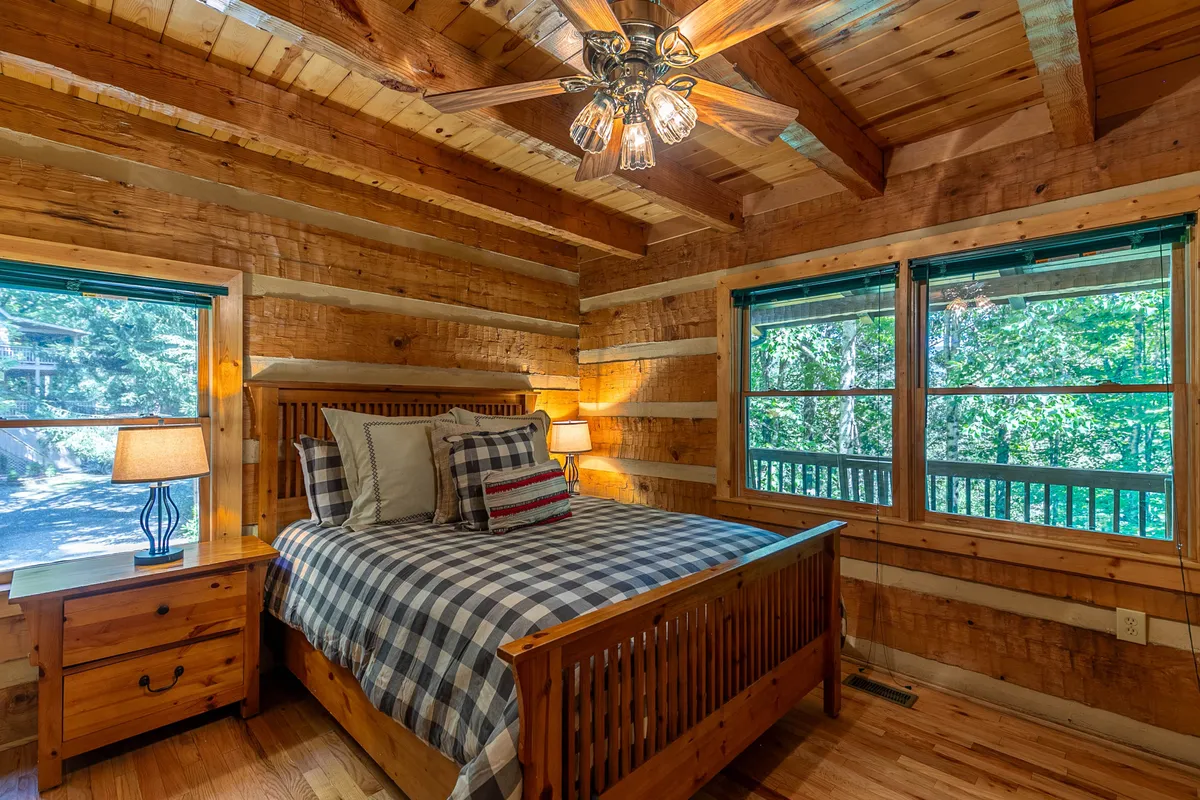 A cozy bedroom with a wooden bed adorned in plaid bedding sits beside windows that offer leafy, nature-filled views. The room is bathed in natural light, creating a warm retreat perfect for relaxation and unwinding after a day outdoors.