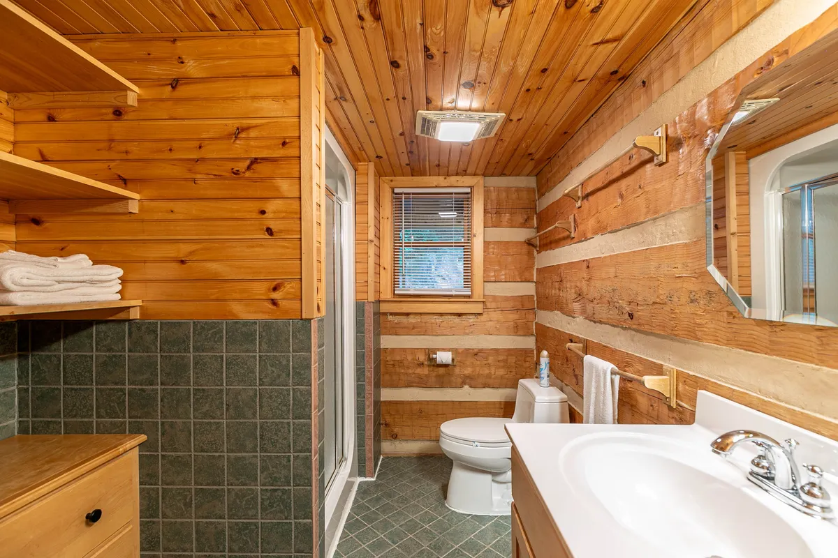 This image features a cozy bathroom with wooden walls, a sink, mirror, shower area, and toilet. The space is warm and inviting, ideal for refreshing after outdoor activities.