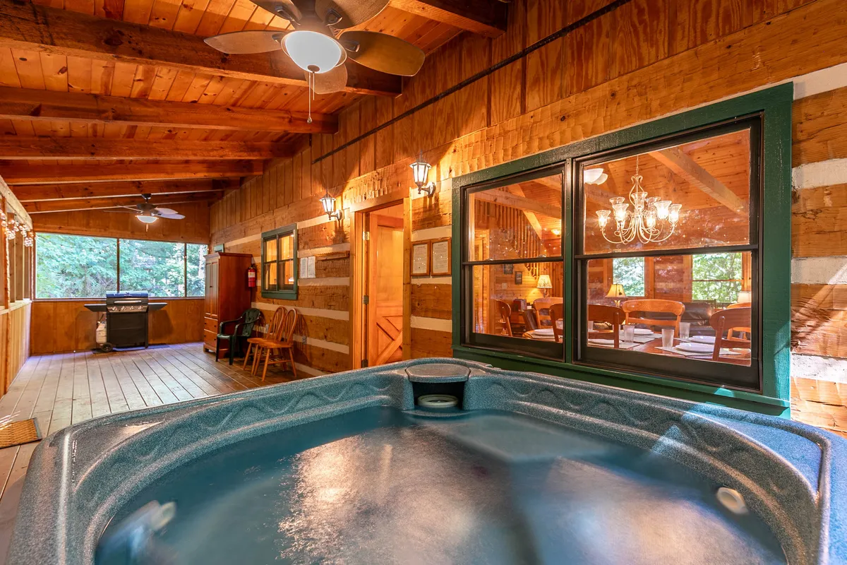 An interior view featuring a private hot tub set against large windows, with wooden beams and a chandelier enhancing the tranquil ambiance. The space is designed for relaxation, with views of the trees and room for gatherings or quiet moments.