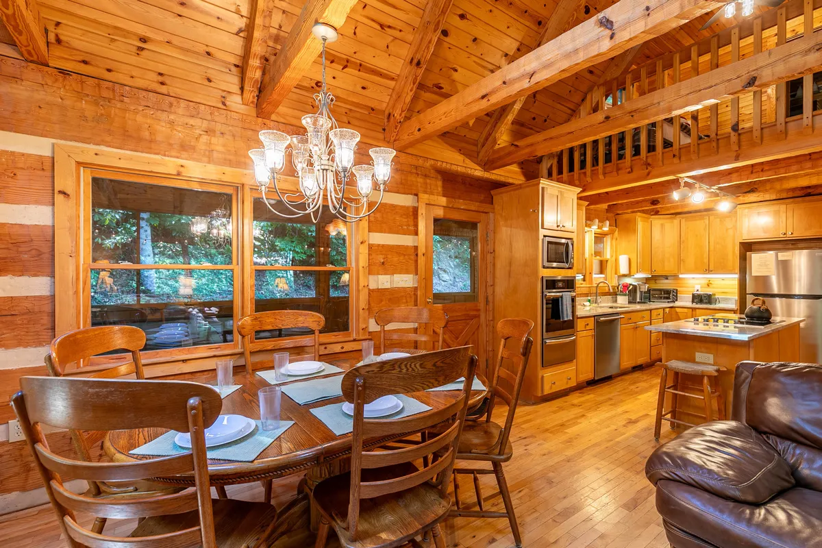 A dining area featuring a wooden dining table with chairs, an elegant chandelier overhead, and a view into a well-appointed kitchen. Visible are the wooden beams and walls that create a rustic yet refined ambiance, with enough space for group meals and celebrations.