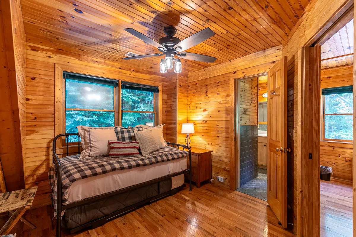 A bed with decorative pillows is illuminated by natural light from windows, accompanied by a wooden bedside lamp. The room exudes a peaceful ambiance, perfect for reading or enjoying quiet moments while appreciating the surrounding scenery.