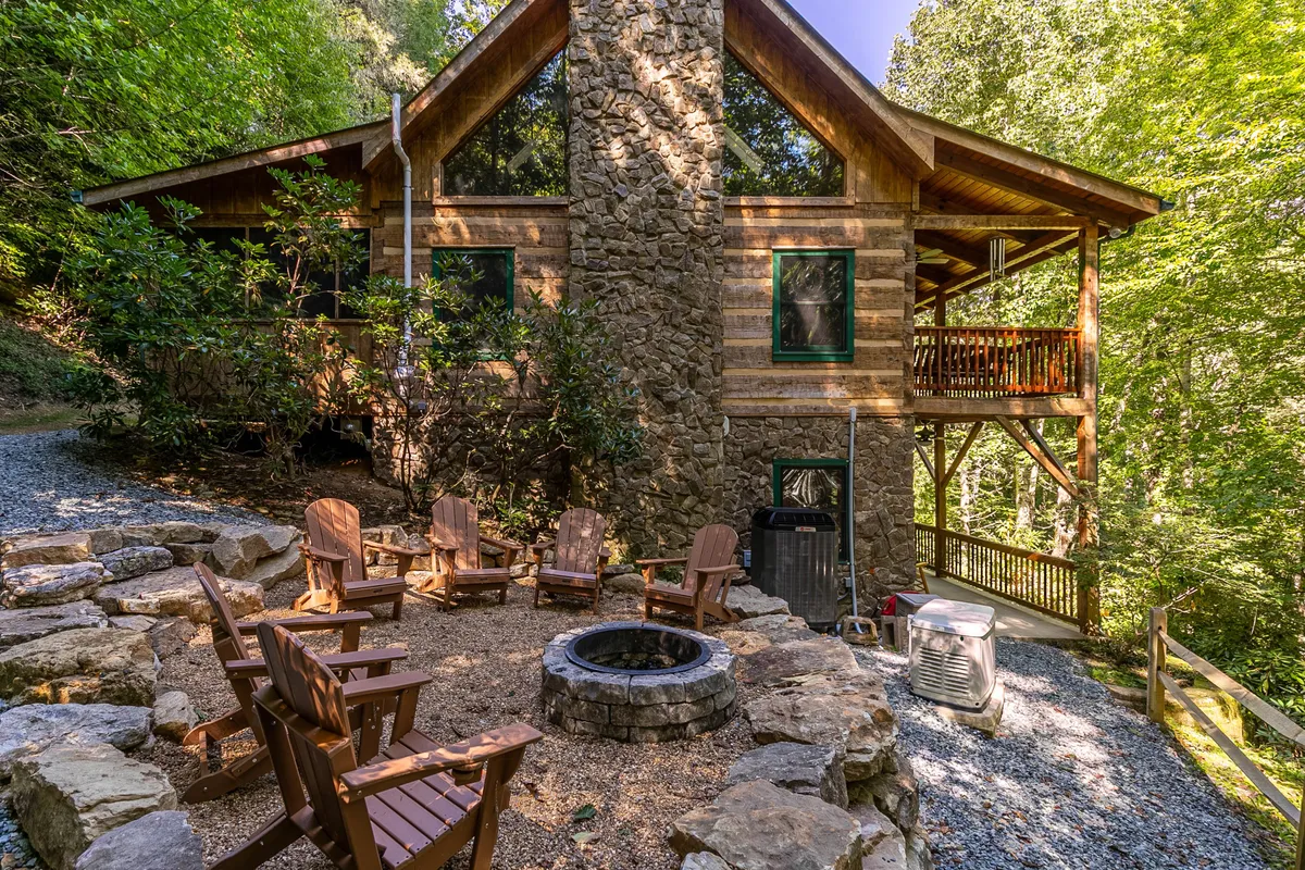 The image showcases the rustic exterior of a cozy log cabin nestled among tall trees, with a prominent stone chimney, a spacious wooden deck, and an inviting fire pit surrounded by Adirondack chairs. The tranquil setting is framed by lush greenery, hinting at a secluded mountain retreat ideal for outdoor gatherings.