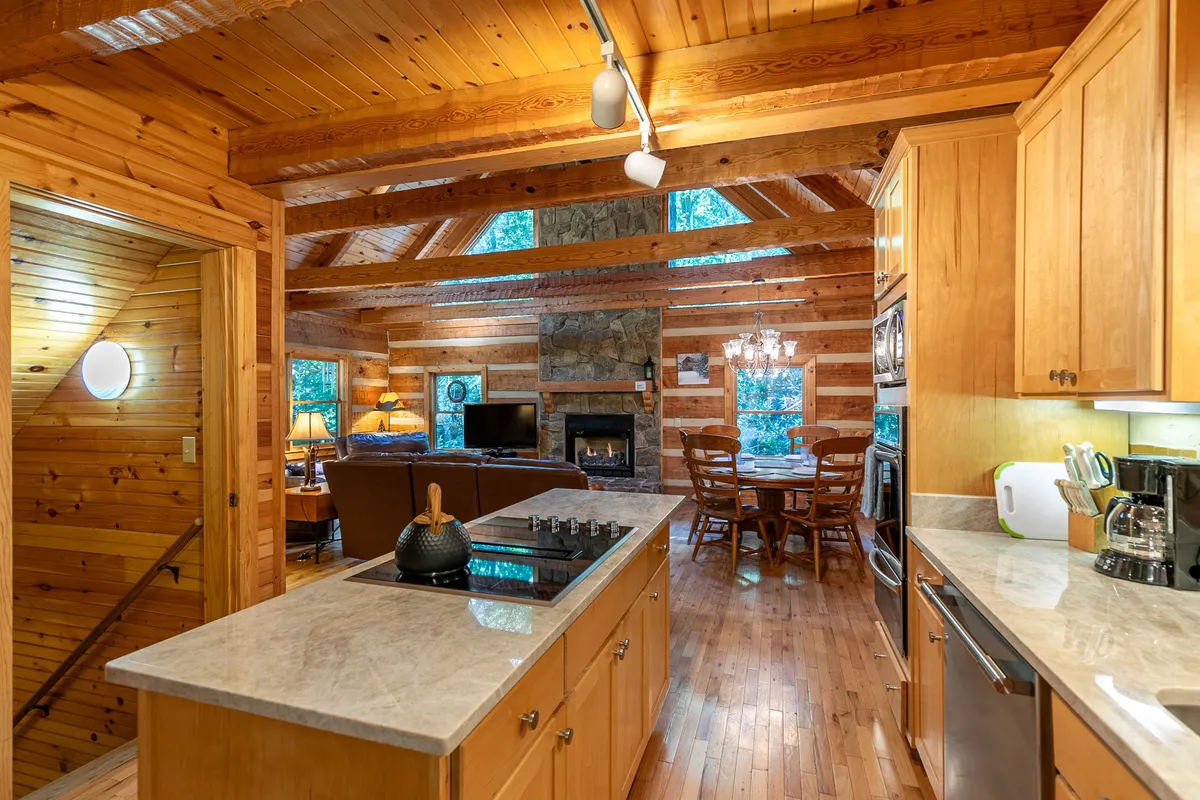 This interior image highlights a kitchen island, fireplace, dining area, and large windows that let in abundant natural light. The space feels open and connected to the outdoors, suggesting a hub for gathering, meal prep, and planning adventures.