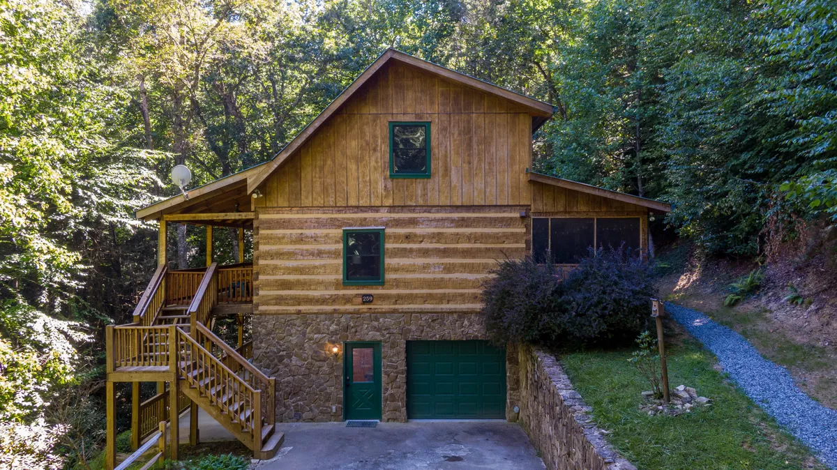 A broad view of the cabin’s exterior, showcasing multiple levels of wooden porches and a stone base, with stairs leading up to inviting outdoor spaces. Hints of activity, such as family gatherings and relaxing on the porch, are suggested by the scene's mood and setup. The lush surroundings and mountain setting are visible, grounding the property firmly in its natural environment.