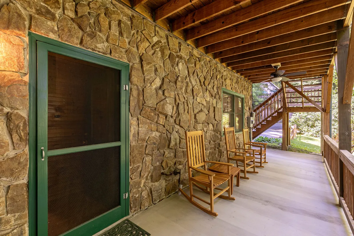 This exterior image showcases a rock wall, wooden rocking chairs on a porch, and a staircase leading up, all surrounded by natural light filtering through the trees. The cozy porch setting looks out to the mountain surroundings, inviting guests to relax and take in the outdoors.