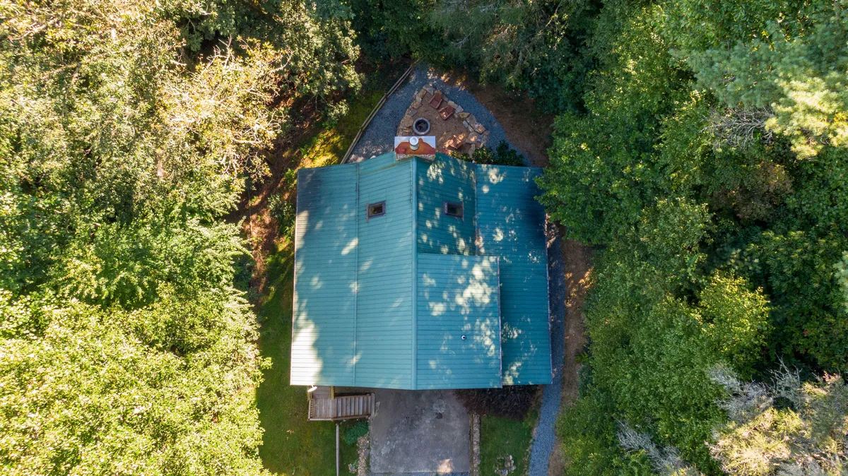 A wide exterior view of Skylark cabin nestled among the trees, with a cozy fire pit area in the foreground and the house’s welcoming porch visible. The setting suggests a peaceful mountain retreat surrounded by nature, perfect for outdoor gatherings and evening storytelling sessions.