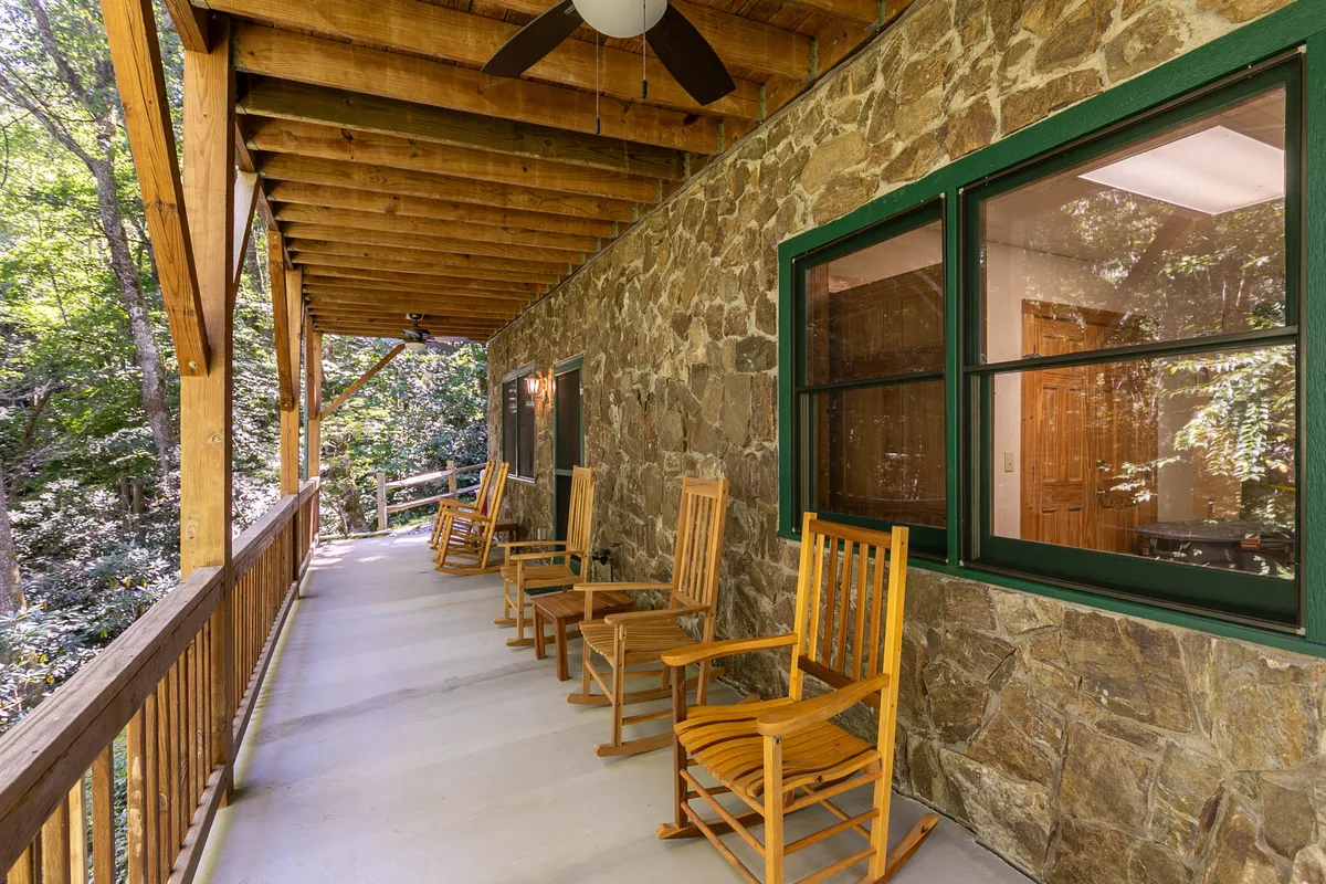 This exterior image captures a cozy porch with wooden rocking chairs, a stone wall, and green-trimmed windows nestled under a wooden ceiling. The seating area is surrounded by nature, inviting guests to relax and savor the mountain ambiance.
