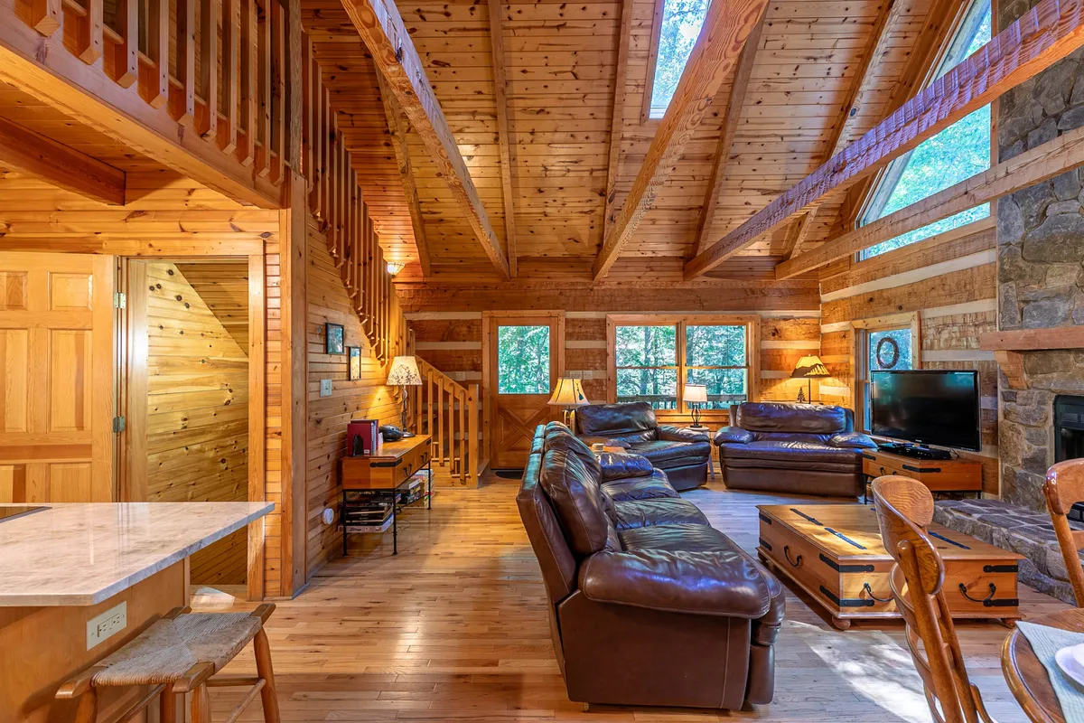 A spacious living area with large windows that allow ample natural light to pour in, accented by rustic wooden beams and a stone fireplace. Leather sofas are arranged invitingly around a coffee table, creating a cozy spot that feels closely connected to the outdoors through the visible views and open feel.