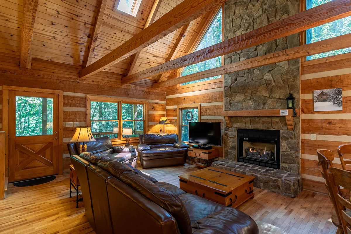 A spacious great room with a striking stone fireplace, large windows that bring in natural light, leather sofas, and exposed wooden beams. The layout invites gathering and relaxation, with the fireplace as a focal point for cozy evenings and socializing.