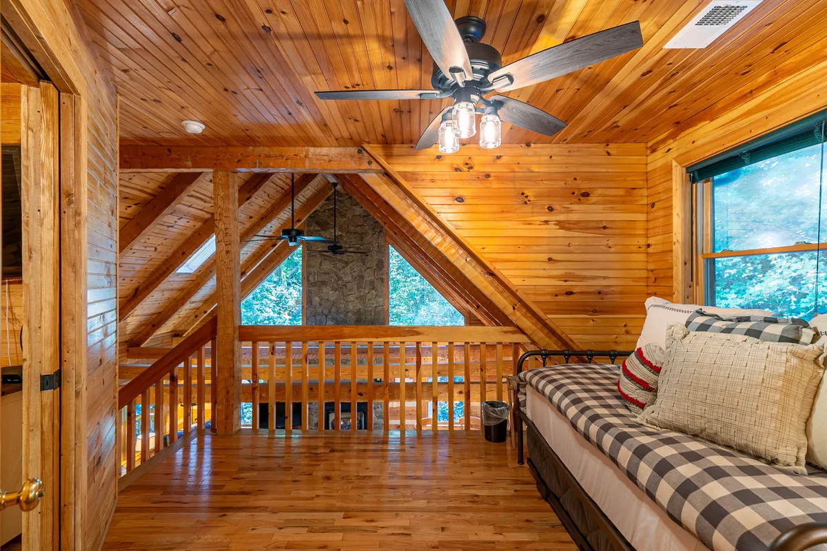 A loft area with a daybed surrounded by cushions sits beneath exposed wooden ceiling beams. Large windows flood the space with daylight, while a cozy stone fireplace anchors the room, inviting relaxation and flexible use for families or friends.