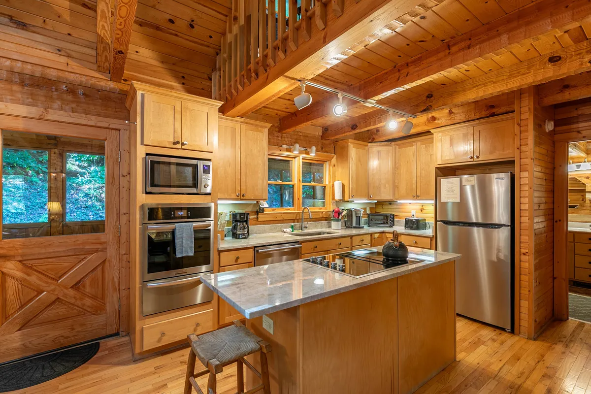 A bright kitchen area with a central island, wooden cabinets, and modern appliances, illuminated by large windows letting in natural light. The scene feels homely and functional.