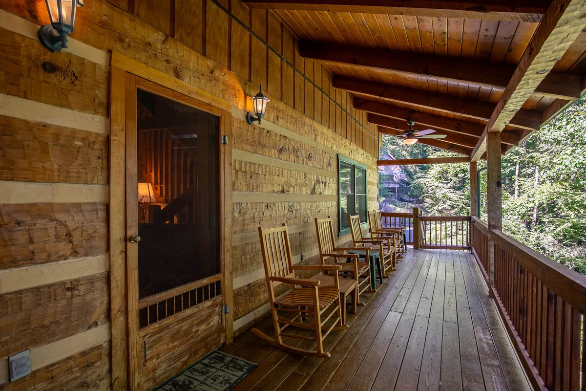 A cozy exterior porch scene featuring several wooden rocking chairs arranged along the log cabin walls. Hanging lanterns add a soft glow, and a screened door hints at easy indoor-outdoor flow. The setting is tranquil, perfect for morning coffee or evening relaxation while enjoying the sounds and sights of nature.